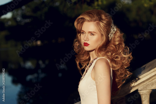 Closeup fashion style portrait of young beautiful woman with golden hair with curls and hairstyling posing outdoors in opark against lake at sunset.