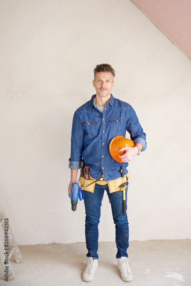 Smiling mature construction man portrait with handyman belt and safety helmet and drill Smiling mature construction man portrait with handyman belt and safety helmet and drill