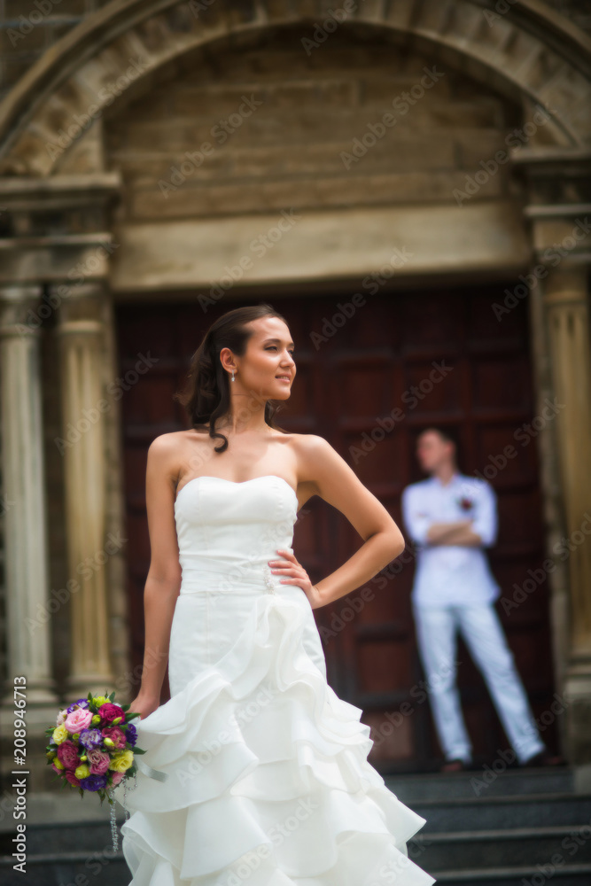 Naklejka premium Wedding photo with the bride and groom. The bride stands epically and looks to the side, and behind her stands the groom near the Catholic Church