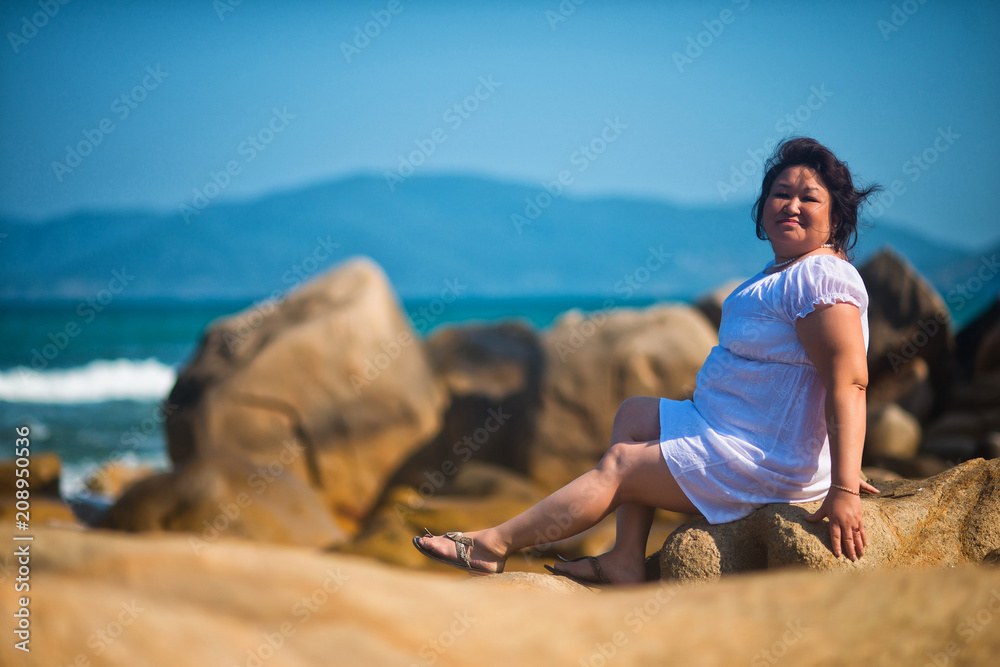 Adult Asian fat woman sitting on the beach in the middle of large boulders on the sea background