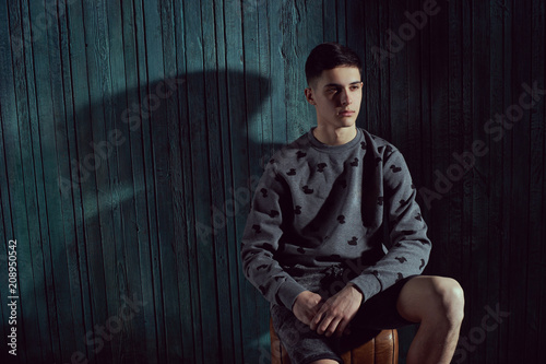 Thoughtful young handsome man with dark short hair sitting against green wooden wall in dark like and looking at you or in camera photo