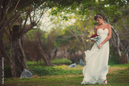 Beautiful young bride posing in a wedding dress in the middle of tropical trees and looks at the wedding bouquet