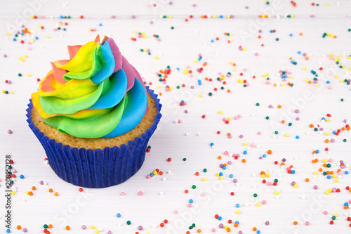 Cupcake with rainbow colorful cream in blue cup on white wooden table decorated with colorful sprinkles.