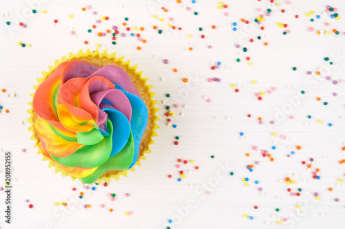 Cupcake with rainbow colorful cream in yellow cup on white wooden table decorated with colorful sprinkles. Top view.