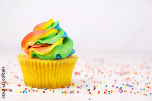 Cupcake with rainbow colorful cream in yellow cup on white wooden table decorated with colorful sprinkles.