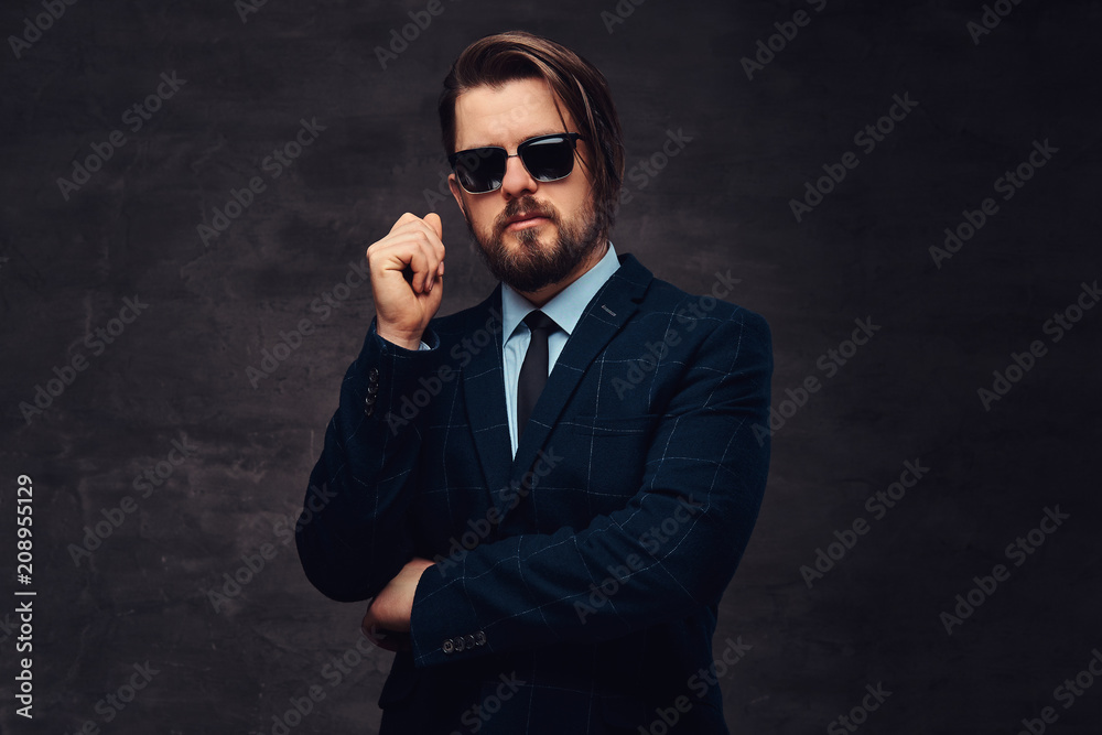 Close-up portrait of a handsome fashionable middle-aged man with beard and hairstyle dressed in an elegant formal suit and sunglasses. Isolated on a textured dark background in studio.