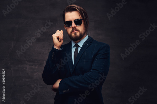 Close-up portrait of a handsome fashionable middle-aged man with beard and hairstyle dressed in an elegant formal suit and sunglasses. Isolated on a textured dark background in studio.