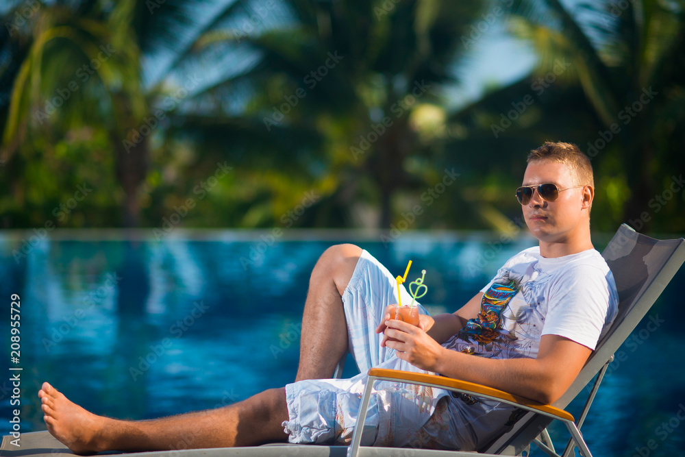 A handsome man in sunglasses lying on a sun lounger by the pool against the palm trees, holding the juice