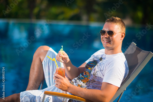 Portrait of a beautiful young man in sunglasses, who lies on a sun lounger near the pool against the palm trees, holding the juice and smiling