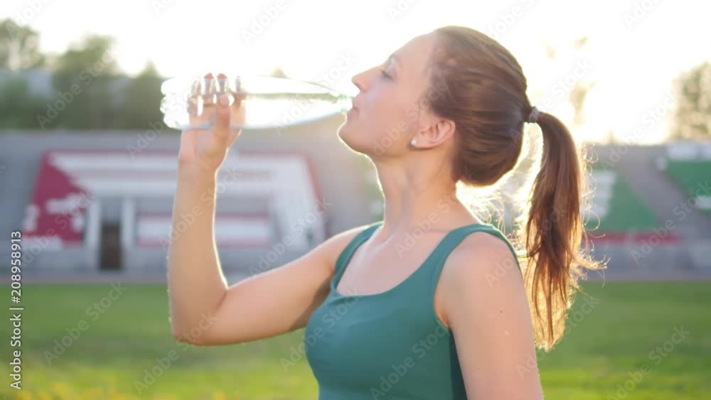 Beautiful young girl tired after jogging drinks water on sunset background Beautiful young girl tired after jogging drinks water on sunset background