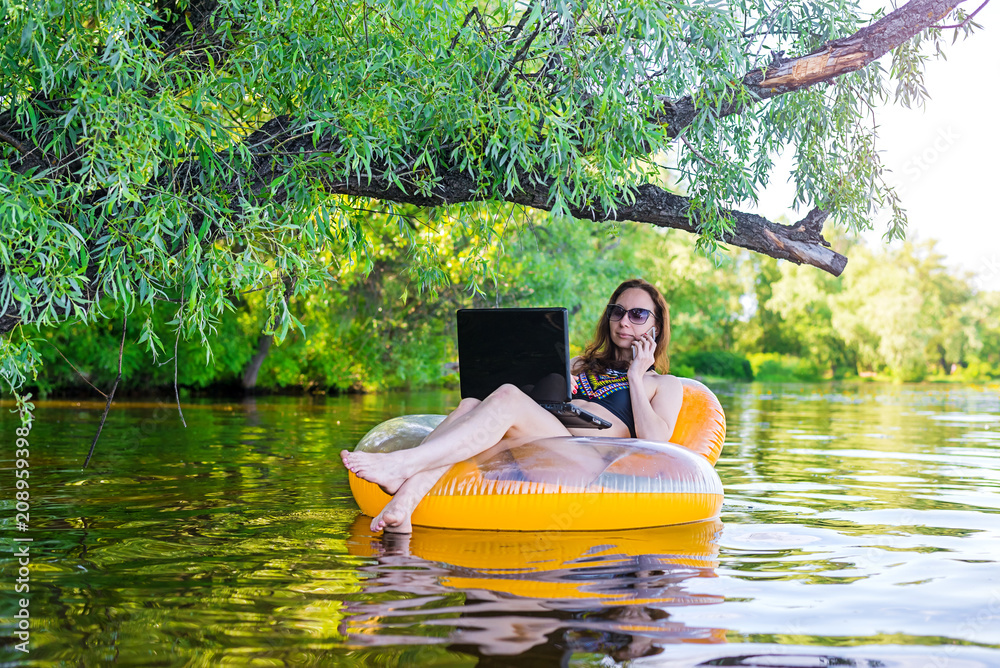 Business woman working on a laptop and talking on smartphone in an inflatable ring in the water, a copy of the free space. Business woman working on a laptop and talking on smartphone in an inflatable ring in the water, a copy of the free space.