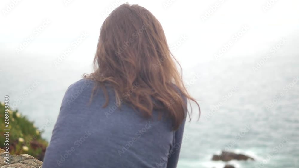 Beautiful young girl looks at the ocean, draws his hands to it. View from the back. Hair fluttering in the wind