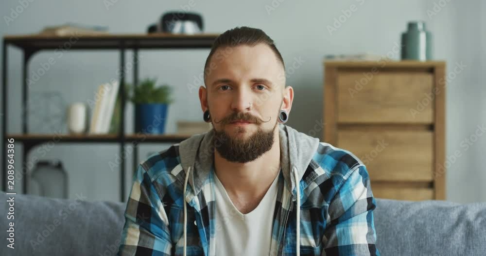 Close up of the hipster man in the motley shirt looking to the camera and shaking no with head. Refuse. Indoor. Close up of the hipster man in the motley shirt looking to the camera and shaking no with head. Refuse. Indoor.