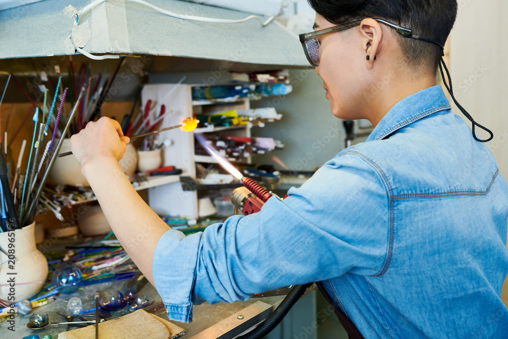 Side view portrait of Asian young woman shaping glass over gas torch while making handmade beads in modern creative workshop, copy space Side view portrait of Asian young woman shaping glass over gas torch while making handmade beads in modern creative workshop, copy space