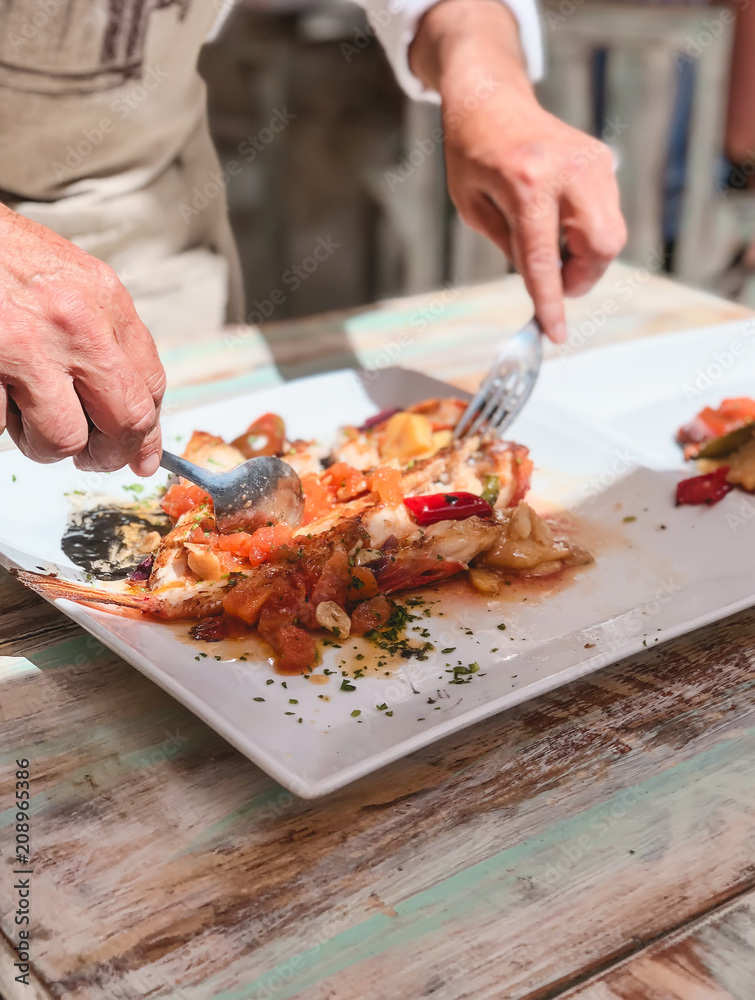 manos de hombre, cocinero de restaurante, preparando y repartiendo pescado tipico de ibiza, en los platos de los comensales, sobre mesa de madera rustica, en el exterior con luz natural de verano