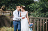 A beautiful bride and a stylish groom stand, embracing forehead to forehead against the backdrop of an old house and a wooden fence, in green foliage. Wedding portrait of walking newlyweds in nature.