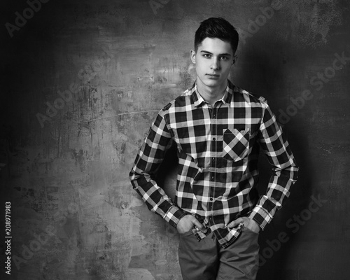 Portrait of young trendy handsome man with short dark hair wearing checkered shirt and brown trousers standing and posing against gray concrete wall