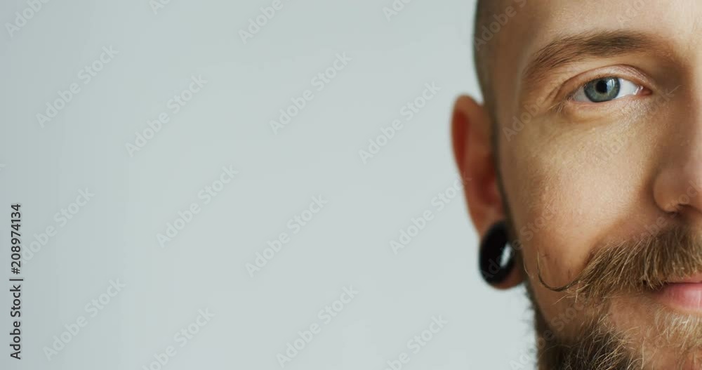 Close up of the attractive male hipster half face with earings smiling and looking at the camera. White wall background. Portrait. Inside.