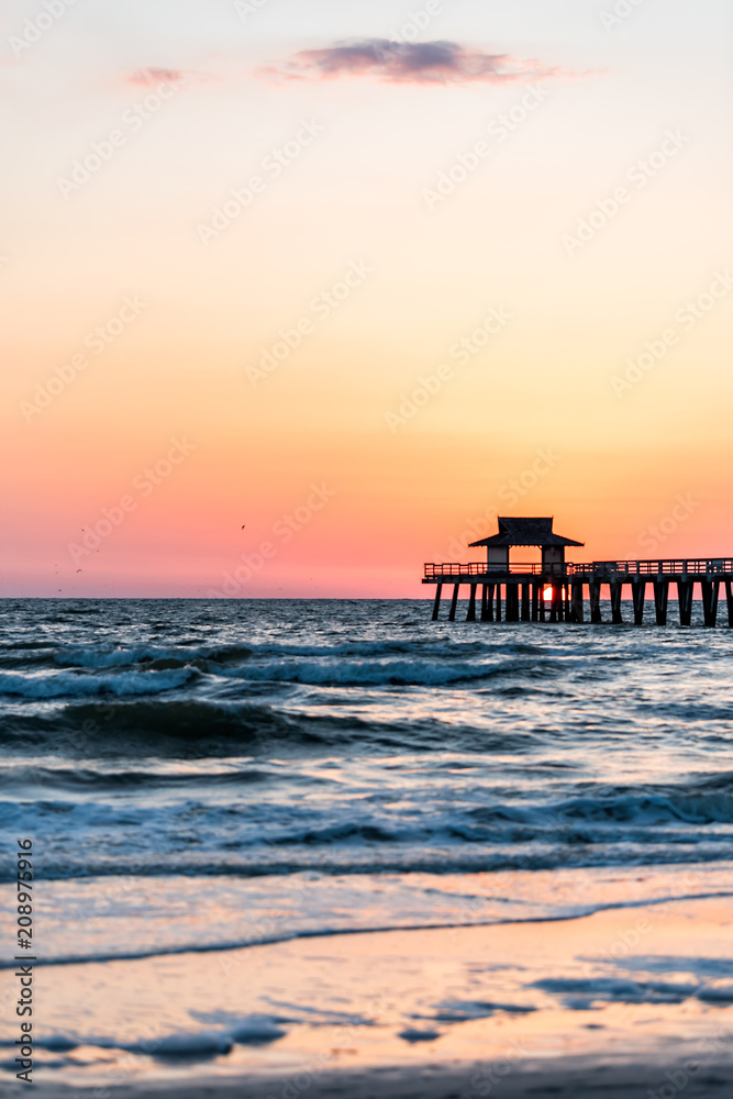 Naples, Florida pink and orange sunset vertical view in gulf of Mexico with sun setting inside Pier wooden jetty, horizon and dark blue ocean waves