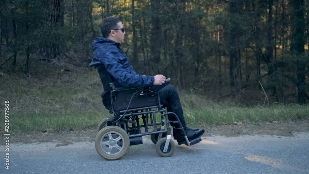 Side view of a handicapped man moving along the road in his powered wheelchair Side view of a handicapped man moving along the road in his powered wheelchair
