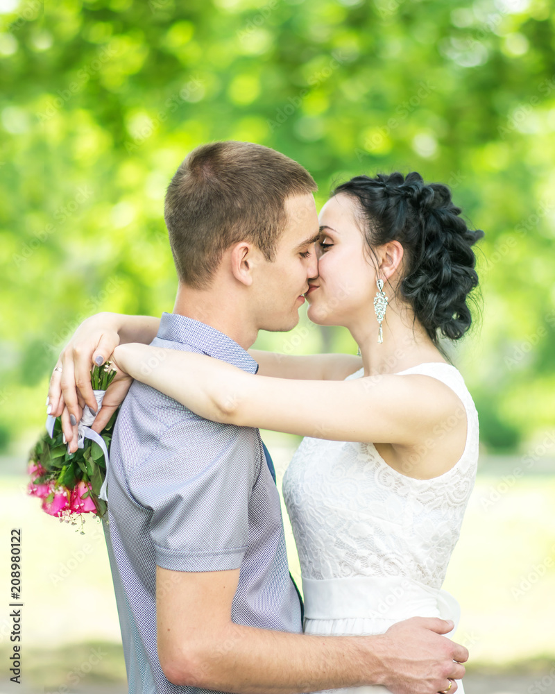 Portrait of beautiful young couple female bride with small wedding pink flowers roses bouquet and male bridegroom kissing in summer park Portrait of beautiful young couple female bride with small wedding pink flowers roses bouquet and male bridegroom kissing in summer park