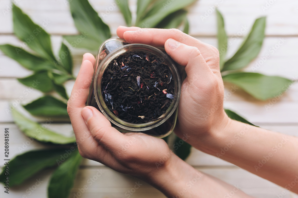 Female hands hold a glass jar with dry tea on a wooden background with twigs and leaves of peonies.