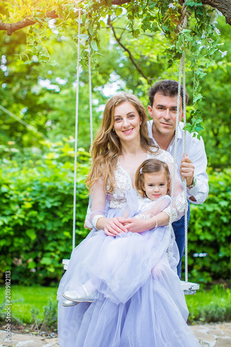 Portrait of happy young family, resting in summer park, mother riding swing in blue dress, riding on swing, and holding small daughter, father is standing behind them
