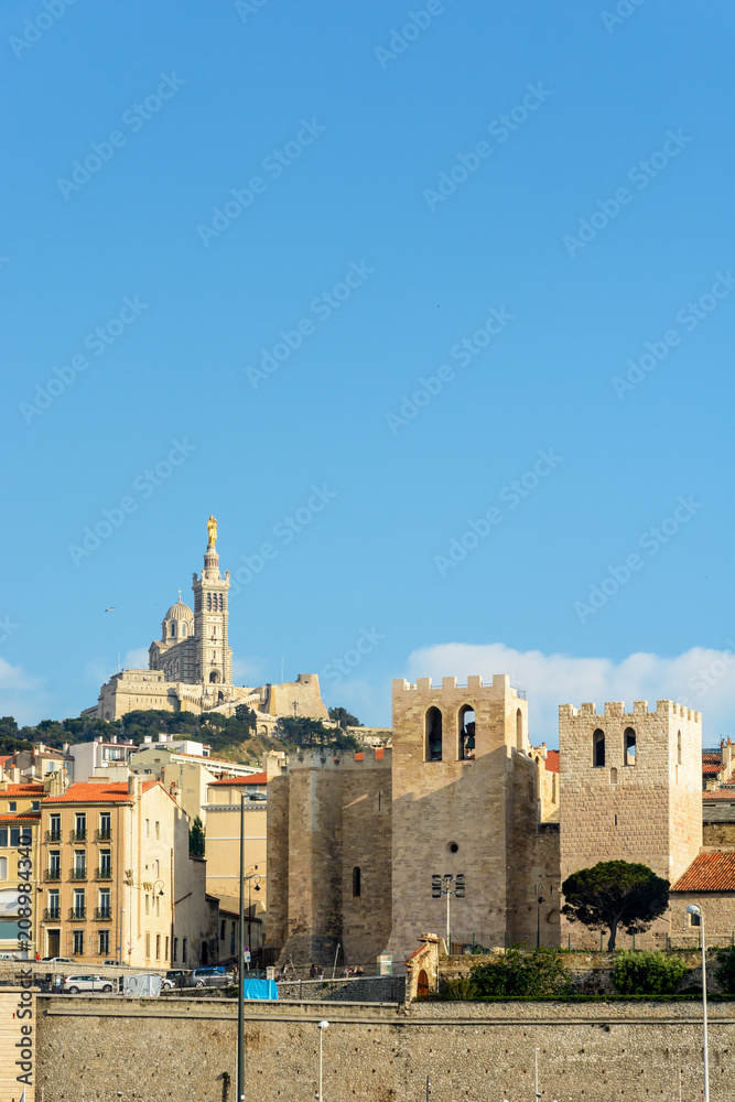 View of the abbey of Saint Victor in Marseille, France, at sunset with its massive fortified square towers, overlooked by the basilica of Notre-Dame de la Garde on top of the hill in the background.