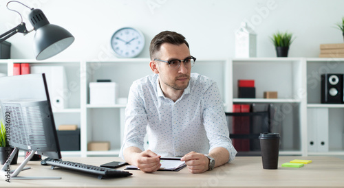 A young man in glasses stands near a table in the office and holds a pencil in his hands.