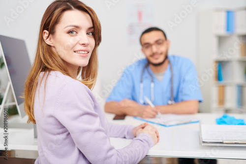 Portrait of beautiful young woman turning to look at camera while sitting at desk in doctors office during consultation, copy space