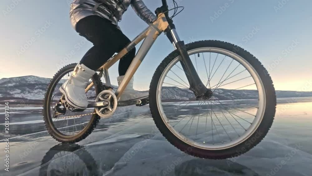 Woman is riding bicycle on the ice. The girl is dressed in a silvery down jacket, cycling backpack and helmet. Ice of the frozen Lake Baikal. The tires on the bicycle are covered with special spikes Woman is riding bicycle on the ice. The girl is dressed in a silvery down jacket, cycling backpack and helmet. Ice of the frozen Lake Baikal. The tires on the bicycle are covered with special spikes