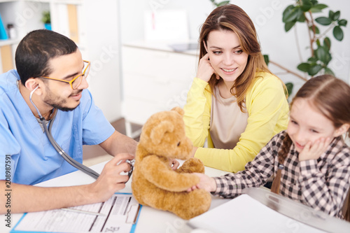 Side view portrait of young Middle-Easter doctor putting stethoscope to plush Teddy Bear toy during consultation with little girl and her mother in modern clinic.