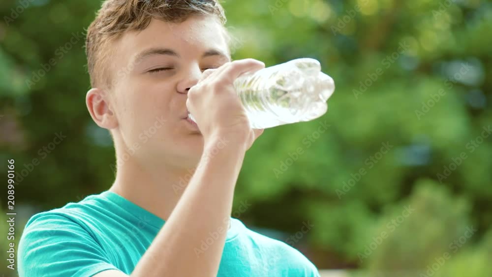 Happy teen boy drinking water from plastic bottle in summer park. Cute teenager drinks cold water - outdoors portrait. Happy teen boy drinking water from plastic bottle in summer park. Cute teenager drinks cold water - outdoors portrait.