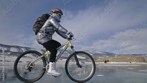 Woman is riding bicycle on the ice. The girl is dressed in a silvery down jacket, cycling backpack and helmet. Ice of the frozen Lake Baikal. The tires on the bicycle are covered with special spikes photo