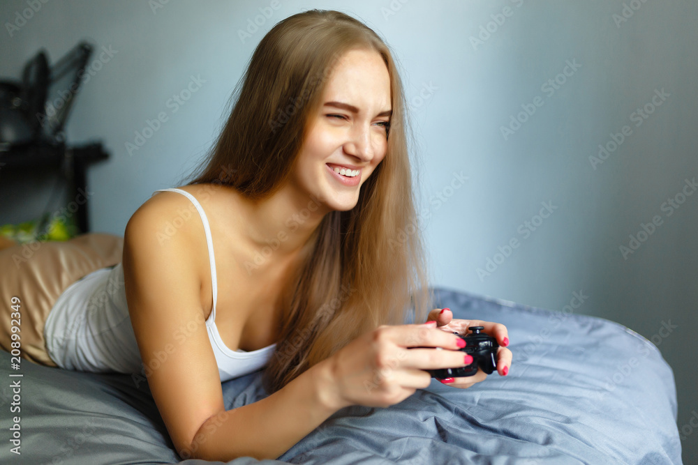 Girl gamer plays with a wireless gamepad while looking at the screen in front of her. Young blonde girl smiles and enjoys winning with gamepad in hand, playing video games console, lying on the bed. Girl gamer plays with a wireless gamepad while looking at the screen in front of her. Young blonde girl smiles and enjoys winning with gamepad in hand, playing video games console, lying on the bed.