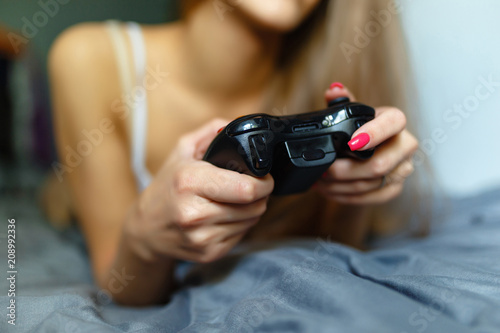 Close-up of hand blonde girl with gamepad in hand playing video games console while lying on the bed. The girl is out of focus, holding the joystick of her video console while playing video games.