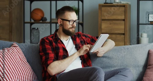 Caucasian male hipster with an earing and in the red motley shirt sitting on the sofa in the living room, taping on the tablet device, thinking and twisting his moustache. Inside