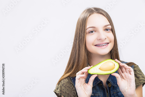 Dental Health Concepts. Positive caucasian Teenager Girl Wearing Teeth Braces Posing with Avocado Against White.