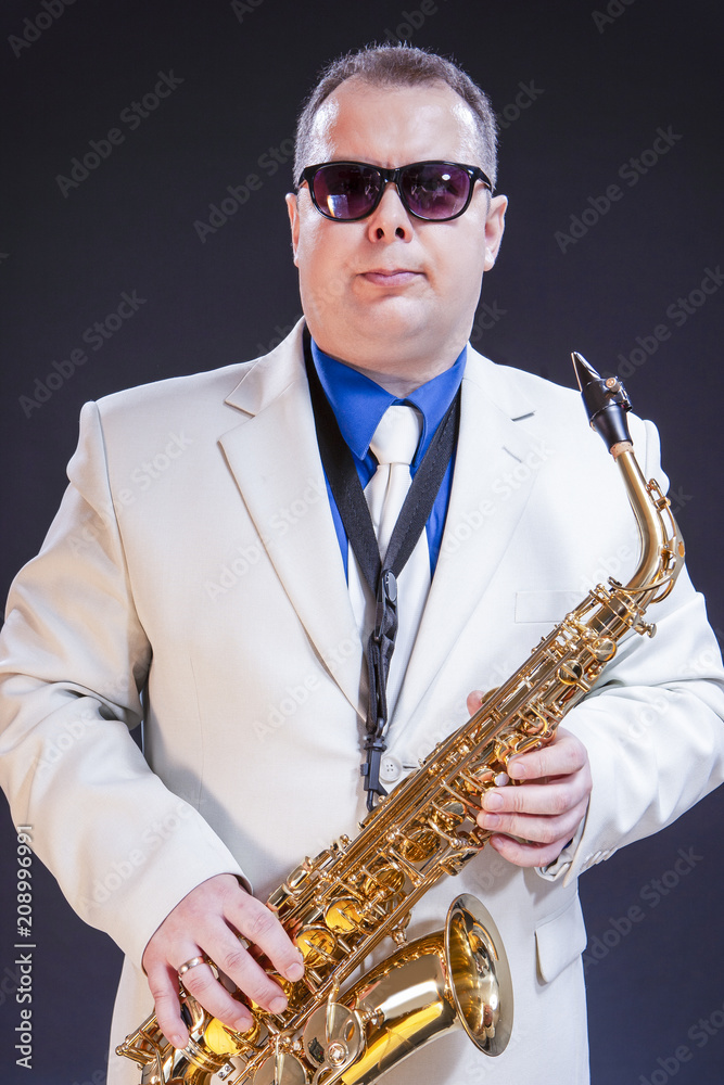 Fototapeta premium Music Themes. Portrait Of Confident Mature Male Saxophonist Posing with Instrument in White Suit and Sunglasses. Against Black Background.