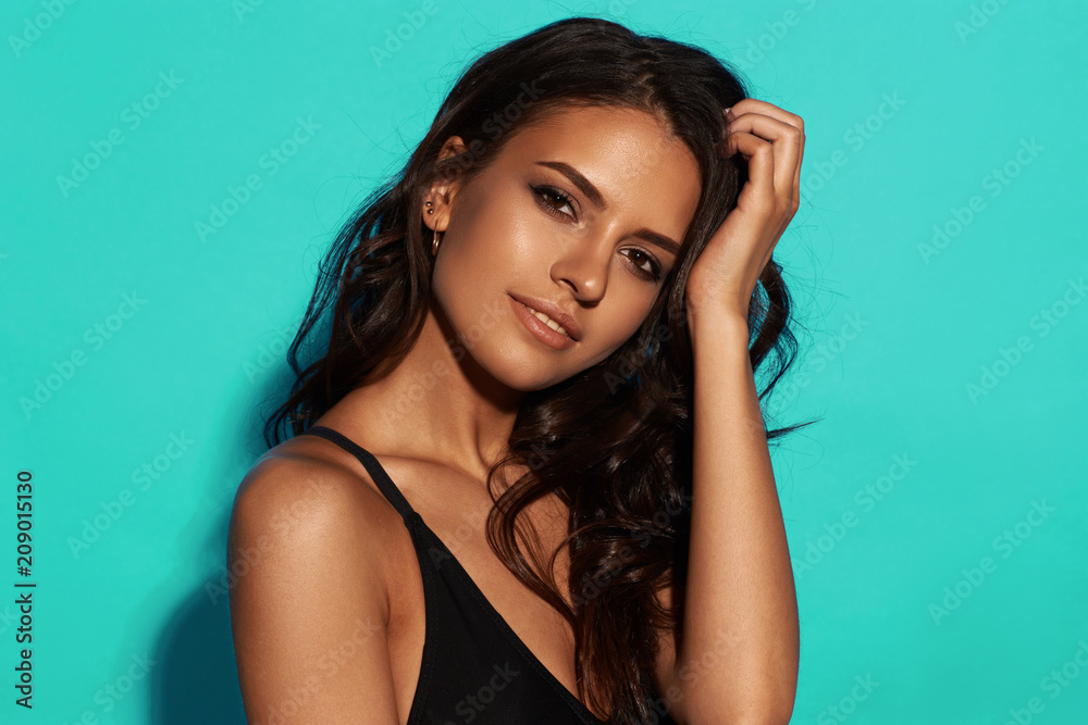 Obraz premium Young sexy slim tanned woman in black swimsuit posing against blue background. Closeup Fashion portrait of beautiful girl with long wavy brunette hair. Swimwear or bikini model
