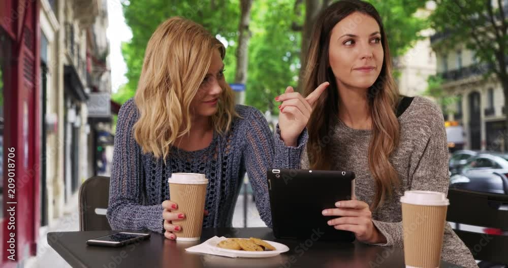 Couple of joyful girls sitting with drinks and looking at tablet in front Paris shops, Two beautiful caucasian woman using portable tablet and socializing on Paris avenue, 4k Couple of joyful girls sitting with drinks and looking at tablet in front Paris shops, Two beautiful caucasian woman using portable tablet and socializing on Paris avenue, 4k
