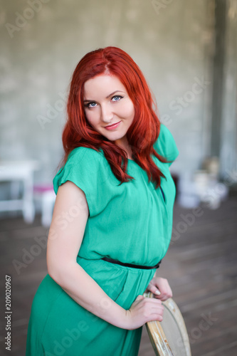 Image of redhead young happy lady in green dress standing over blurred grey wall background looking camera