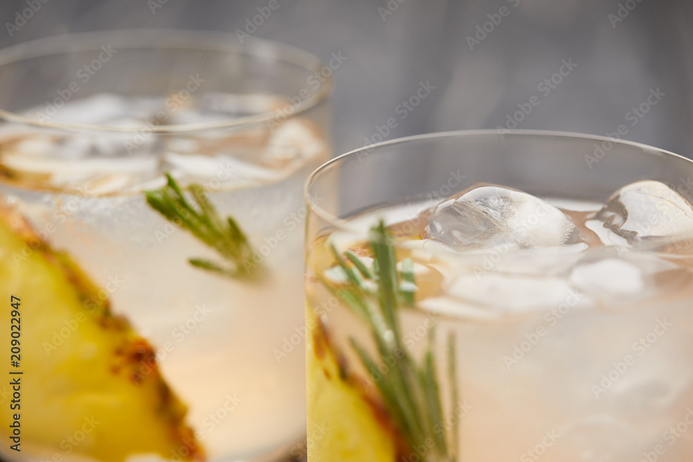 selective focus of two glasses of lemonade with pineapple pieces, ice cubes and rosemary on grey wooden tabletop selective focus of two glasses of lemonade with pineapple pieces, ice cubes and rosemary on grey wooden tabletop