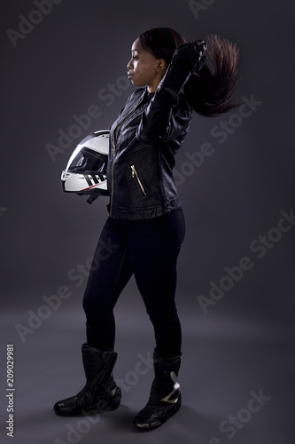 Black female motorcycle biker or race car driver or stuntwoman wearing leather racing suit and holding a protective helmet. She is standing confidently in a studio