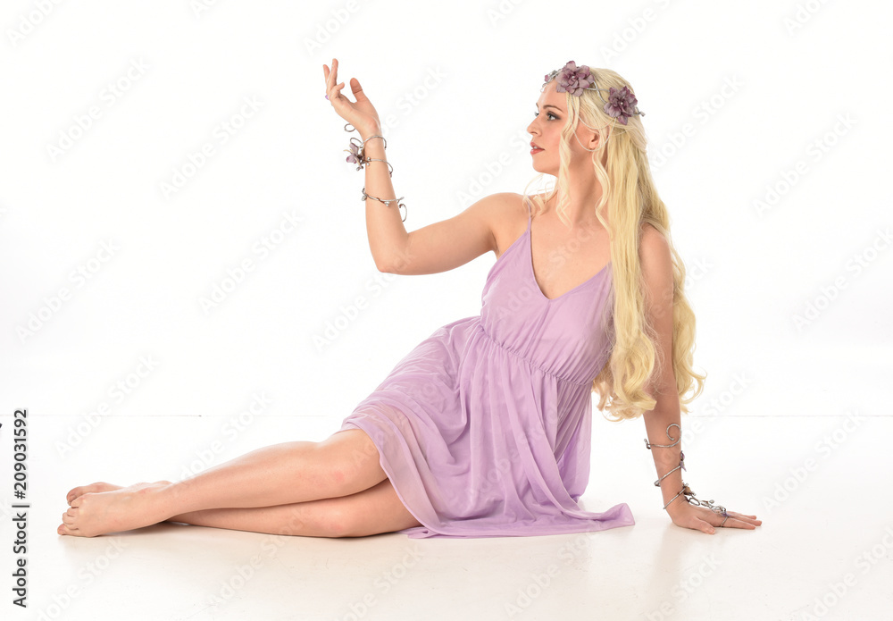 full length portrait of a blonde fairy girl ,wearing purple dress. seated pose on white studio background.