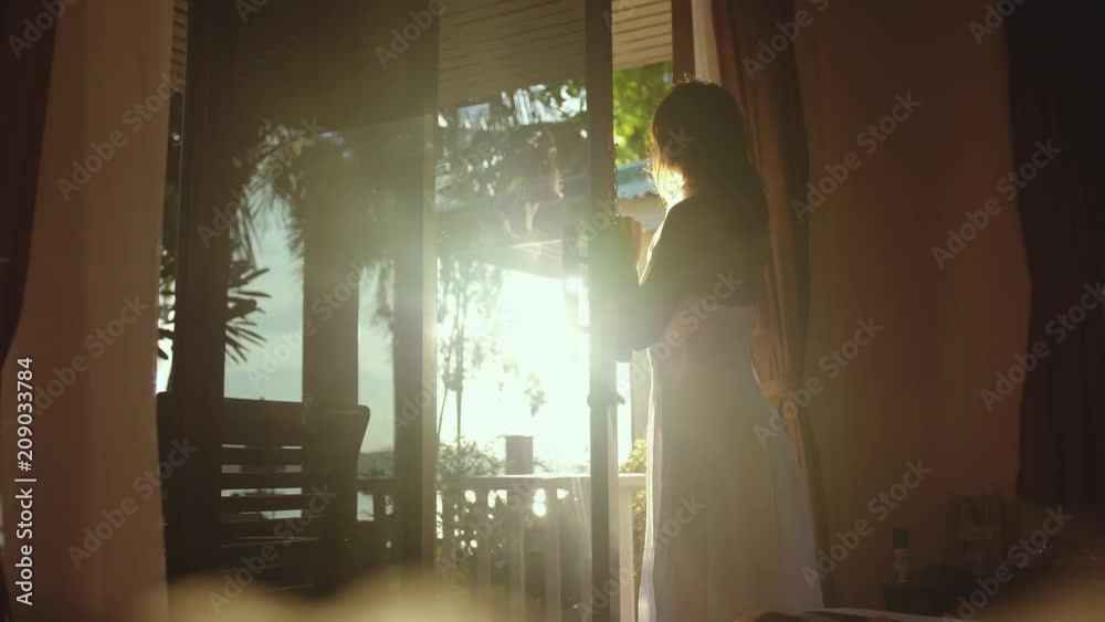 Pretty girl opens the door in the early morning during sunrise with lens flare effects and go out to the terrace. slow motion. 3840x2160
