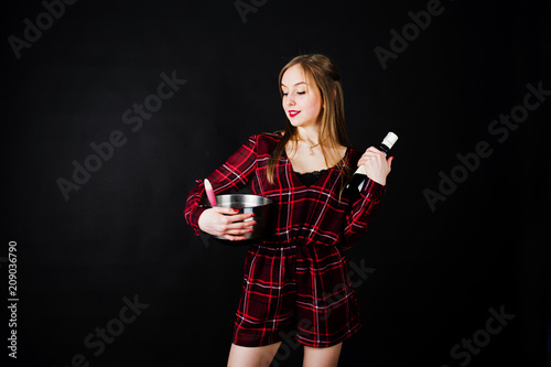 Young housewife in checkered dress with saucepan and kitchen spoon and bottle isolated on black background.