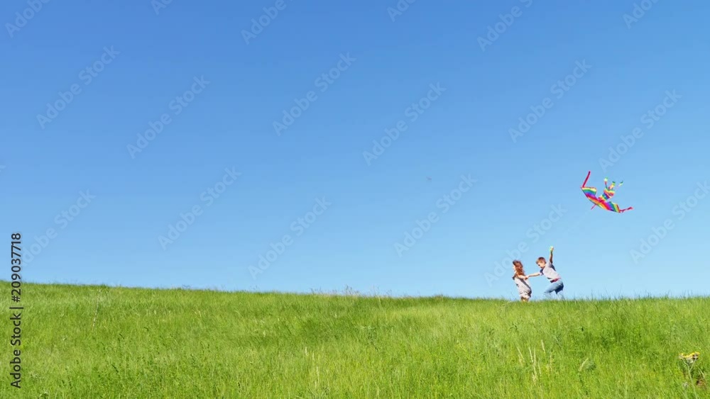 Happy childhood - Children run along the top of the green hill with a flying kite at clear blue sky background. Wide shot, slow motion Happy childhood - Children run along the top of the green hill with a flying kite at clear blue sky background. Wide shot, slow motion
