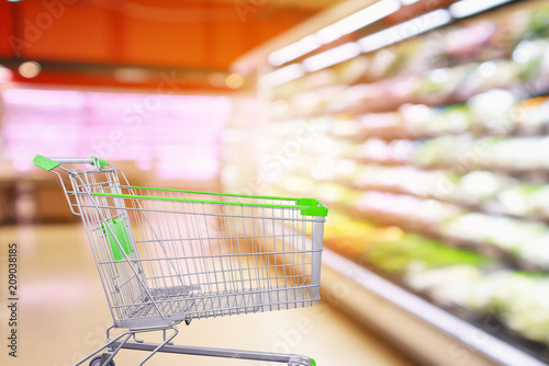 Supermarket aisle with fresh vegetables and fruit shelves interior blur background with empty shopping cart