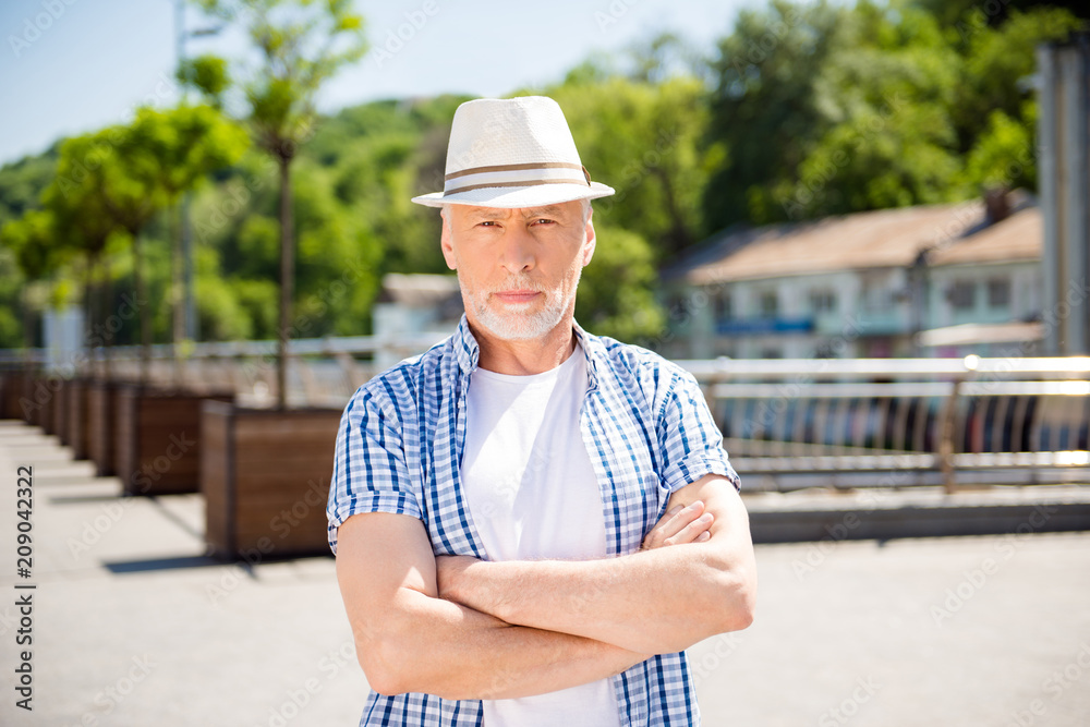 Portrait of concentrated dreamy businessman in straw hat holding arms crossed looking at camera standing over blurred street background